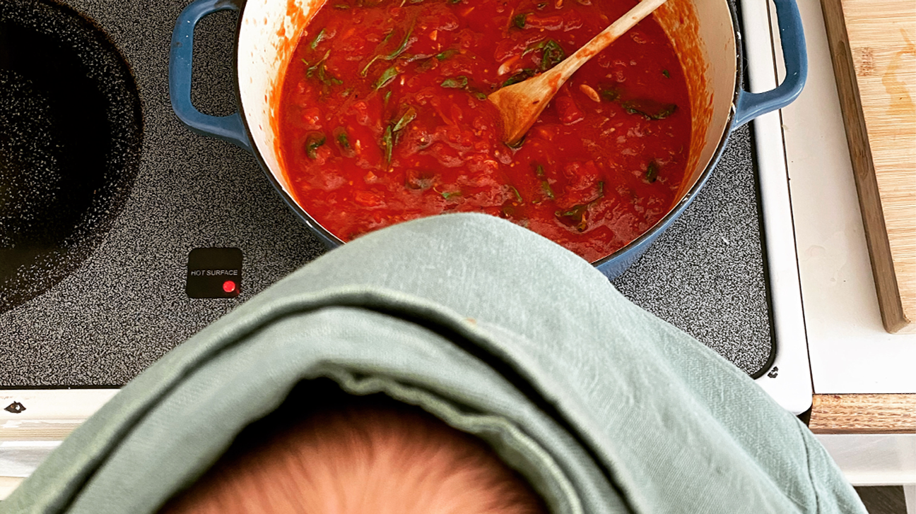 photo from the POV of a mom looking down at a pot of red sauce on the stove with her sleeping baby strapped to her front