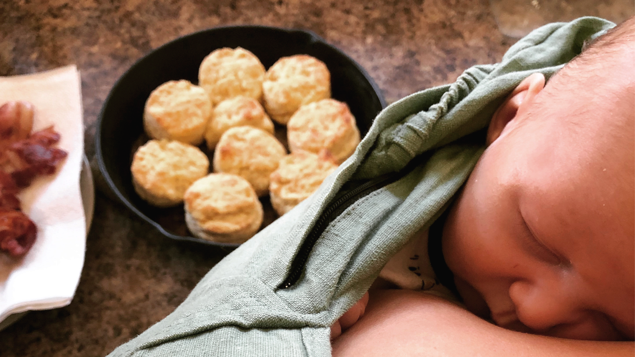 a photo from the POV of a mom looking down at a pan of biscuits with her sleeping baby wrapped securely to her front